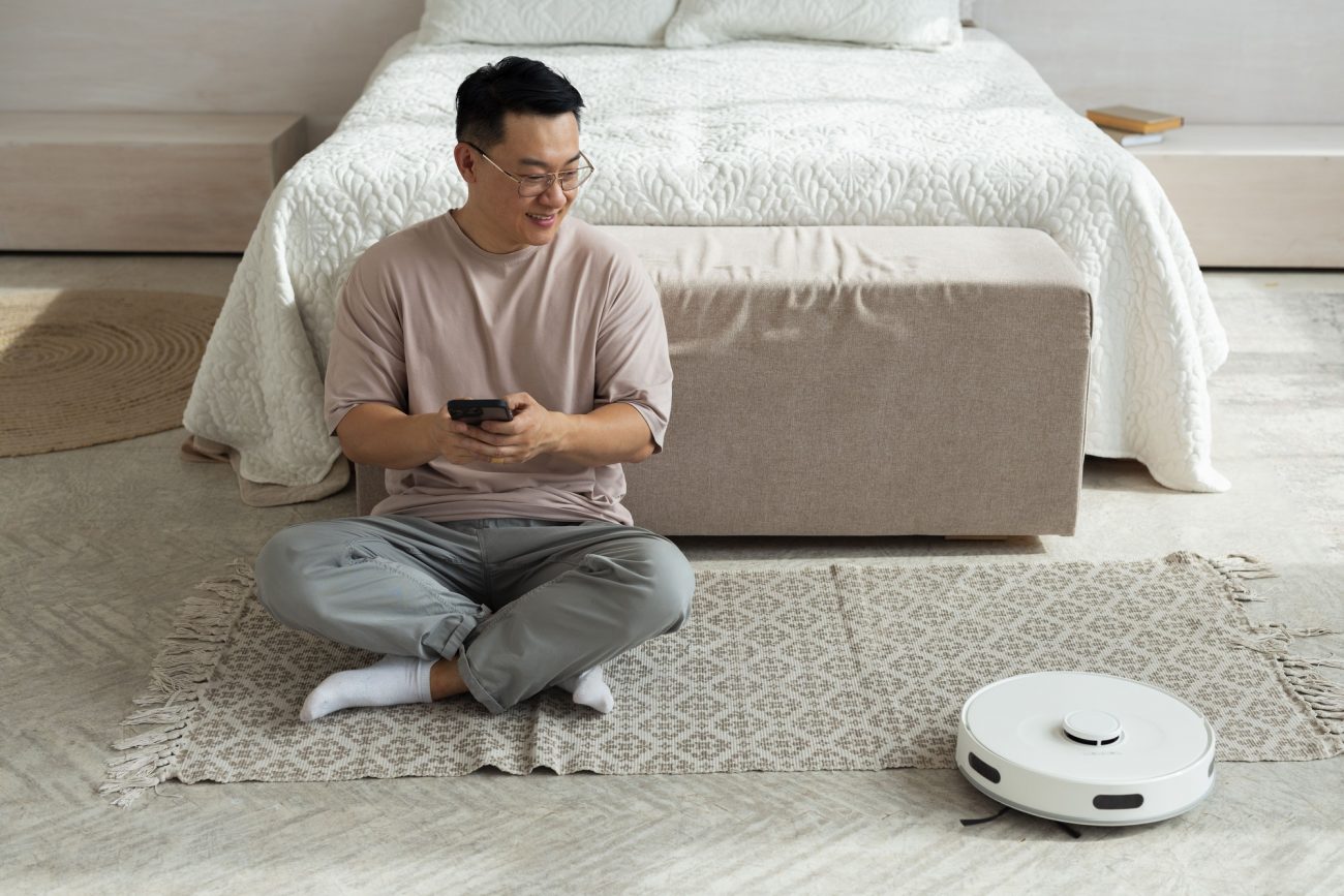 Siurblys robotas su šerelių šepečiais. Vyras sėdi ant grindų ir su telefonu nustato roboto nustatymus.