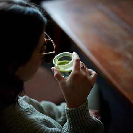 Ceremoninė Matcha arbata permatomoje stiklinėje rankoje